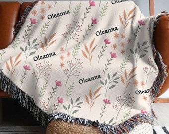May include: Cream-colored throw blanket with a floral pattern of pink flowers, green leaves, and brown foliage. The word "Oleanna" is printed across the blanket. The blanket has a fringed edge in black, white, and other colors.