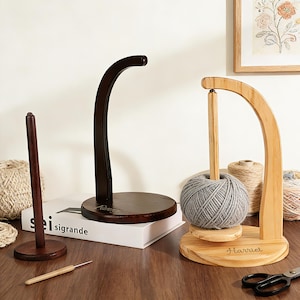 May include: Wooden yarn holders in various styles. One is dark brown with a curved arm, another is natural wood with a gray yarn ball, and a third is a simple spindle. A crochet hook and scissors are also present.