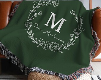 Pode incluir: Um cobertor verde escuro com um design de coroa de flores branca e a letra "M" no centro. As palavras "The Morlains" estão escritas abaixo. O cobertor tem bordas com franjas.