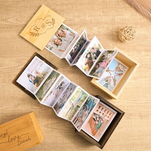 May include: Wooden photo boxes with personalised initials and names, containing accordion-style photo albums. The albums display multiple photos, showcasing memories. The boxes are light brown and dark brown, with a natural wood grain finish.