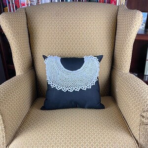 May include: A gold armchair with a black pillow featuring a white crocheted collar. The chair is in front of a bookcase filled with books.