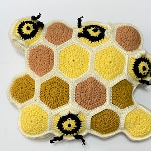 Può includere: Un cuscino a nido d'ape all'uncinetto con quattro api ricamate. Il nido d'ape è fatto di filato bianco, giallo, marrone e dorato. Le api hanno strisce nere e gialle e occhi neri.