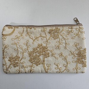May include: A rectangular zippered pouch made of off-white fabric featuring an intricate gold thread embroidery pattern of floral clusters and winding vines. The pouch includes a metallic zipper closure along the top edge with a small metal pull.