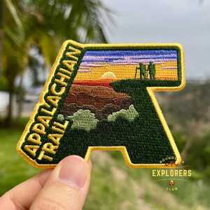 Può includere: Toppa ricamata raffigurante l'Appalachian Trail. La toppa ha la forma della lettera "A" con una scena paesaggistica di un tramonto ed escursionisti. Il testo "APPALACHIAN TRAIL" è arcuato sul lato sinistro. L'angolo in basso a destra recita "EXPLORERS CLUB".