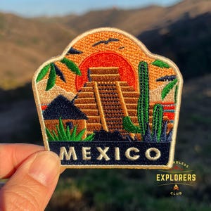 Könnte beinhalten: Gestickter Patch mit einer Sonnenuntergangsszene über einer Maya-Pyramide, mit Kakteen und Palmen. Der Patch ist beige mit einem blauen Rand und dem Wort "MEXICO" in weißen Buchstaben. Der Patch stammt vom "Endless Explorers Club".