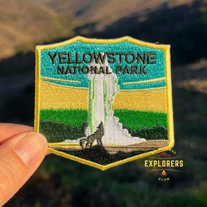 May include: Embroidered patch featuring Yellowstone National Park, with a green, yellow and blue background. The patch depicts a geyser erupting with a wolf in the foreground. The text "Yellowstone National Park" is in black lettering. The patch has a yellow border and a small "Explorers Club" logo in the bottom right corner.