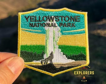 Op de afbeelding: Gestikte patch met het Yellowstone National Park, met een groene, gele en blauwe achtergrond. De patch toont een geiser die uitbarst met een wolf op de voorgrond. De tekst "Yellowstone National Park" staat in zwarte letters. De patch heeft een gele rand en een klein "Explorers Club" logo rechtsonder.
