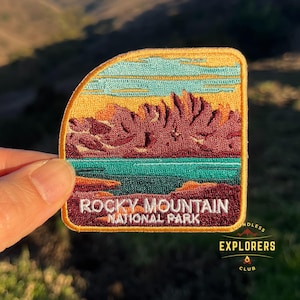 May include: An embroidered patch depicting a mountain range with a lake in the foreground. The text "Rocky Mountain National Park" is displayed in white on a burgundy background.