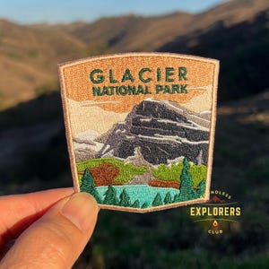 Pode incluir: Um remendo bordado nas cores marrom e bege com o texto "Glacier National Park" e uma representação de uma cordilheira e um lago. O remendo tem uma borda branca e o texto "Endless Explorers Club" no canto inferior direito.