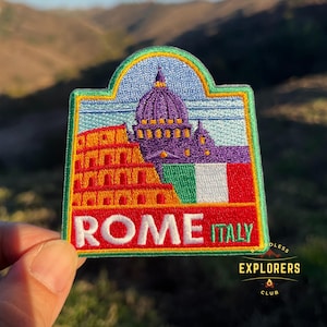 Puede incluir: Un parche bordado colorido que representa el Coliseo y la Basílica de San Pedro en Roma, Italia. El parche tiene un borde verde y el texto "ROMA ITALIA" en letras blancas sobre un fondo rojo.