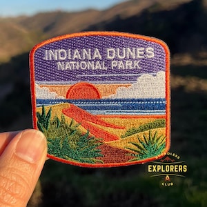 May include: An embroidered patch featuring a sunset scene over a beach and dunes. The patch is purple, orange, yellow, and blue. The text "Indiana Dunes National Park" is embroidered in white on the patch.