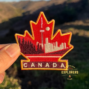 以下が含まれることがあります： 赤いと白い刺繍の入ったパッチで、メープルリーフの形をしています。街のシルエットと「CANADA」という文字が黄色で書かれています。パッチは黄色の縁取りがあり、「ENDLESS EXPLORERS CLUB」という文字が黒で書かれています。