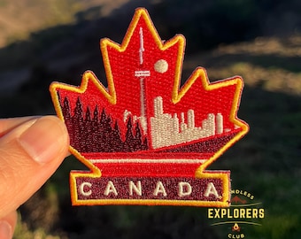 Puede incluir: Parche bordado rojo y blanco con forma de hoja de arce con una silueta de ciudad y la palabra "CANADA" en letras amarillas. El parche tiene un borde amarillo y el texto "ENDLESS EXPLORERS CLUB" en letras negras.