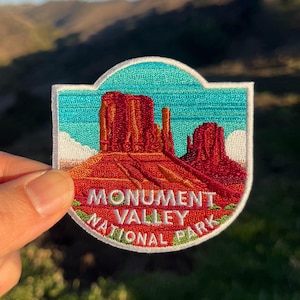 Può includere: Una toppa ricamata bianca con una catena montuosa rossa e arancione su uno sfondo di cielo blu. Il testo "Monument Valley National Park" è ricamato in bianco su uno sfondo verde.