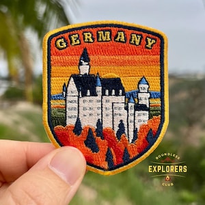 Può includere: Toppa ricamata raffigurante una scena di castello con la scritta "GERMANY" in lettere arancioni. La toppa ha un bordo giallo e un logo che dice "BOUNDLESS EXPLORERS CLUB". Il castello è bianco con accenti blu e neri, su uno sfondo di cielo arancione e blu.