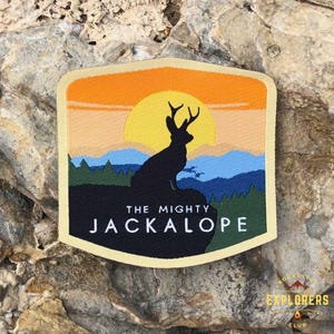 Op de afbeelding: Een beige geborduurde patch met een silhouet van een jackalope met geweien tegen een zonsondergangachtergrond. De tekst "THE MIGHTY JACKALOPE" is in wit op een zwarte achtergrond gedrukt.