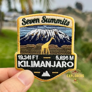 Puede incluir: Parche bordado con un paisaje de montaña con el texto "Seven Summits" y "Kilimanjaro". El parche incluye la altura de la montaña en pies (19.341 FT) y metros (5.895 M), con una silueta de jirafa.