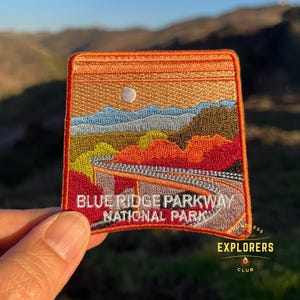 May include: An embroidered patch featuring a scenic view of the Blue Ridge Parkway National Park. The patch depicts a winding road through a mountainous landscape with a sunset sky and a white moon. The text "Blue Ridge Parkway National Park" is embroidered in white on the bottom of the patch.