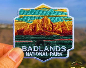 Op de afbeelding: Een witte, blauwe en groene geborduurde patch met de woorden "Badlands National Park" en een afbeelding van een zonsondergang boven een bergketen.