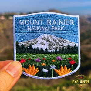 Pode incluir: Um remendo bordado azul e branco com o texto "Mount Rainier National Park". O remendo apresenta um pico de montanha nevado com árvores verdes e um campo de flores coloridas.