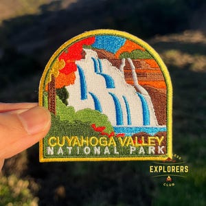 May include: Embroidered patch featuring a waterfall scene with blue water cascading down a white cliff. The patch has a yellow border and text that reads "CUYAHOGA VALLEY NATIONAL PARK" and "EXPLORERS CLUB". The patch includes green, orange, and red foliage.