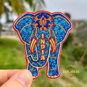 Peut inclure: Écusson brodé en forme d'éléphant avec le mot "INDIA" au centre. L'écusson a un fond bleu avec des détails orange et rouges. L'écusson fait partie du "Boundless Explorers Club".