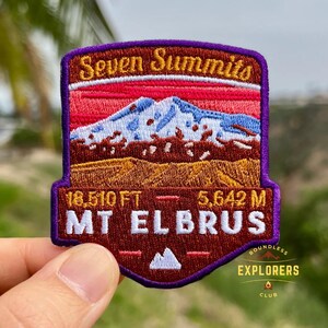 May include: Embroidered patch with the text "Seven Summits" and "MT ELBRUS". The patch features a mountain range design with red, blue, and brown colours. The patch also includes the text "18,510 FT - 5,642 M" and "BOUNDLESS EXPLORERS CLUB".