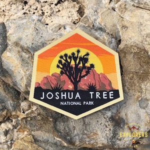 May include: A hexagonal patch with a sunset scene featuring a Joshua Tree silhouette. The patch is beige with orange and red accents. The text "Joshua Tree National Park" is printed in black.