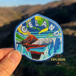 Puede incluir: Un parche bordado azul y blanco con la palabra "ICELAND" en letras blancas sobre un fondo azul. El parche presenta un paisaje colorido con un volcán, montañas y un río. La parte inferior del parche tiene el texto "BOUNDLESS EXPLORERS CLUB" en letras blancas sobre un fondo negro.