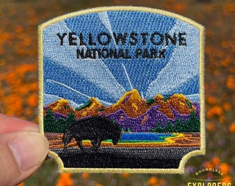 May include: Yellowstone National Park embroidered patch with a blue sky, mountains, and a lake. The patch has a yellow border and a black silhouette of a bison.