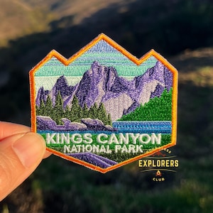 May include: An embroidered patch featuring a mountain range with a lake in the foreground. The patch is orange, blue, green, and purple. The text "Kings Canyon National Park" is on the patch. The text "Explorers Club" is below the patch.