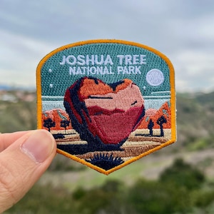 Könnte beinhalten: Ein blauer und gelber gestickter Aufnäher mit dem Text "Joshua Tree National Park" und einer herzförmigen Felsformation in der Mitte. Der Aufnäher hat einen weißen Mond und Sterne im Hintergrund.