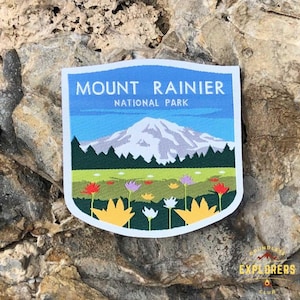 Puede incluir: Un parche bordado azul y blanco con el texto "Mount Rainier National Park". El parche presenta una imagen estilizada del Monte Rainier con un pico nevado y un bosque verde en primer plano. Hay flores coloridas en el campo debajo de la montaña.