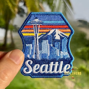 May include: Embroidered patch featuring the Seattle skyline, including the Space Needle, mountains, and city buildings. The patch has a blue border and the word "Seattle" in white script. The background shows a sunset with orange, yellow, and red hues.