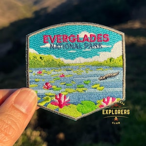 May include: An embroidered patch depicting a scenic view of Everglades National Park. The patch is blue, green and pink with the text "Everglades National Park" in white. The patch is surrounded by a grey border and has the text "Explorers Club" in the bottom right corner.