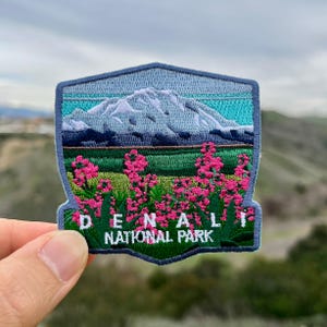 May include: An embroidered patch featuring a mountain range with a blue sky and pink flowers. The text "Denali National Park" is embroidered in white on a green background.