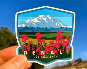 May include: A sticker featuring a blue, green, and white illustration of Denali National Park with a mountain range and pink flowers. The text "Denali National Park" is printed in white on a blue background.