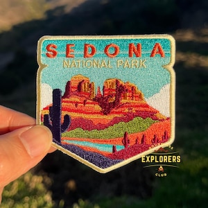 Peut inclure: Un écusson brodé coloré avec le texte "Sedona National Park" et une représentation de formations rocheuses rouges, un cactus et une rivière. L'écusson a une bordure dorée et un petit logo en bas à droite qui dit "Ground Explorers Club".