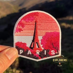 May include: Embroidered iron-on patch featuring a pink and red sunset scene with the Eiffel Tower in the center. The text "PARIS" is below the tower. The patch has a white border and a white background. The text "EXPLORERS CLUB" is in the lower right corner.