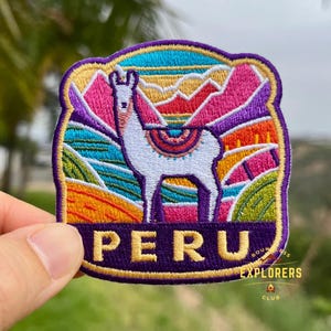 May include: Embroidered patch featuring a white llama with a colorful saddle, set against a backdrop of mountains and fields. The word "PERU" is displayed in gold letters. The patch is bordered in gold and purple, with the words "BOUNLESS EXPLORERS CLUB" in the bottom right corner.