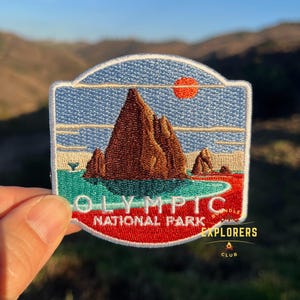 Puede incluir: Un parche bordado con una cordillera de montañas con un sol rojo en el cielo. El parche es rojo, blanco, azul, marrón y verde. El texto "Olympic National Park" está en el parche. El texto "Bundle Explorers Club" está en el parche.