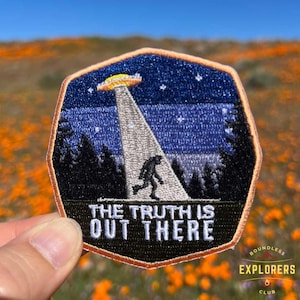 Puede incluir: Parche bordado con un fondo de cielo nocturno azul oscuro, un OVNI, una figura de Bigfoot y el texto "The truth is out there". El parche está rodeado por un borde marrón y tiene un pequeño logotipo de "Boundless Explorers Club" en la esquina inferior derecha.