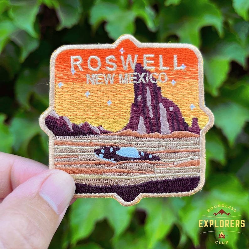 May include: Embroidered patch with a sunset scene of a mountain range and a flying saucer. The patch is orange, brown, and beige with the text "Roswell, New Mexico" in black.