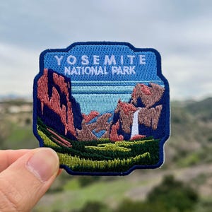 May include: A blue and white embroidered patch with the text "Yosemite National Park". The patch features a scenic view of a mountain range with a waterfall.