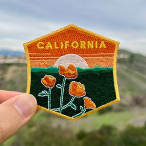 May include: A yellow and orange embroidered patch with the word "CALIFORNIA" in yellow lettering. The patch depicts a sunset with a white sun and green hills in the background. Four orange poppy flowers are in the foreground.