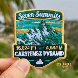 May include: Embroidered patch with the text "Seven Summits" and "Carstensz Pyramid." The patch features a mountain range design in green, gold, and teal, with the height in feet and meters. The patch is from the "Explorers Club."