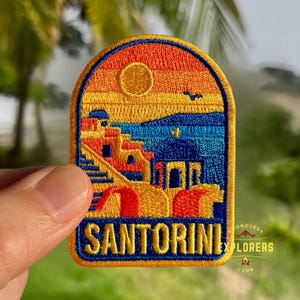Può includere: Toppa ricamata raffigurante un paesaggio di Santorini con un tramonto blu e arancione, edifici e la parola "SANTORINI" in bianco. La toppa ha un bordo blu e il testo "ENDLESS EXPLORERS CLUB".