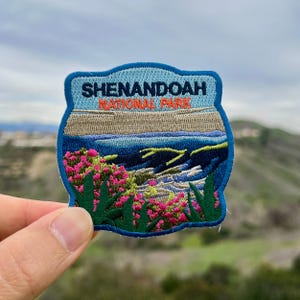 Może przedstawiać: Naszywka haftowana w kolorze niebieskim i zielonym z tekstem "Shenandoah National Park" i panoramicznym widokiem na pasmo górskie z różowymi kwiatami na pierwszym planie.