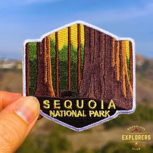 Könnte beinhalten: Gestickter Aufnäher mit einer Darstellung des Sequoia-Nationalparks. Der Aufnäher zeigt Mammutbäume in Braun, Grün und Gelb, mit dem Schriftzug "SEQUOIA NATIONAL PARK" in goldfarbenen Buchstaben. Der Aufnäher wird vor einem verschwommenen Hintergrund gehalten.