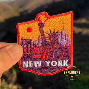 May include: An embroidered patch with a purple and orange cityscape of New York City, including the Statue of Liberty and the Brooklyn Bridge. The patch has a red border and the text "NEW YORK" in white. The bottom right corner of the patch has the text "BOUNDLESS EXPLORERS CLUB" in white.